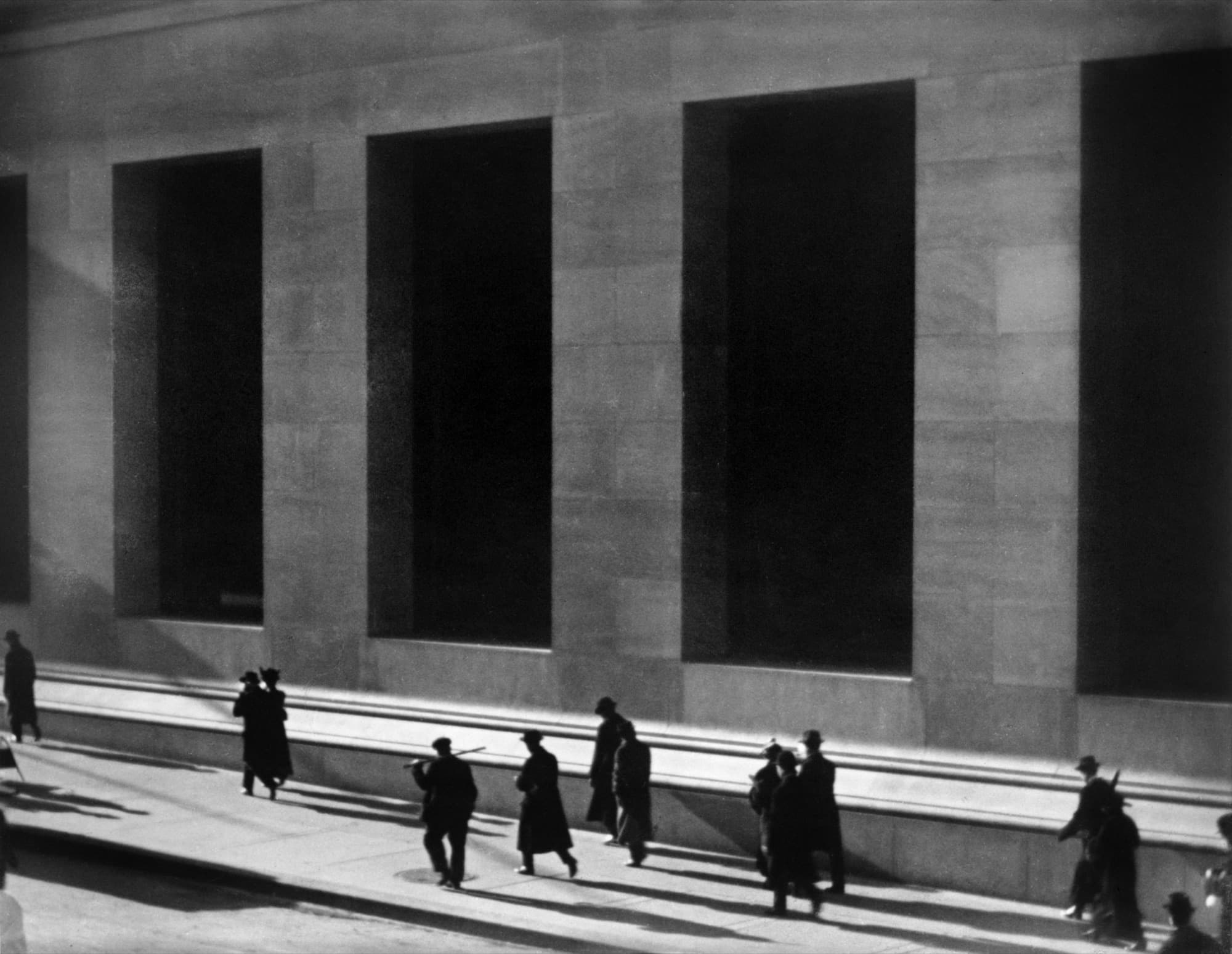 Black-and-white photograph of people walking past the tall dark windows of a Wall Street bank in low morning sun.